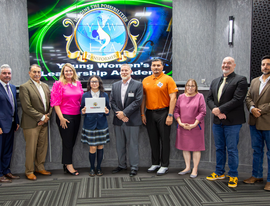 A group of ten people pose indoors in business and casual attire, with a student holding a certificate. A screen behind them displays the Young Womens Leadership Academy logo.
