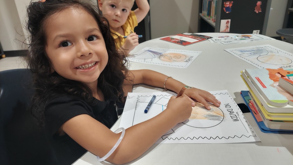 A young student sits at a classroom table smiling while coloring an educational worksheet with a marker. Another young student is seated in the background holding a writing utensil. The table has several student drawings, worksheets, books, and classroom materials. The classroom setting supports hands-on learning and creative activities.