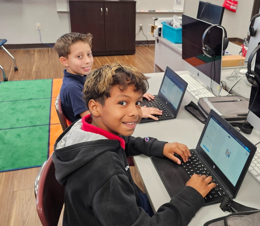 Two young boys sitting at a white desk in a computer lab, smiling warmly at the camera. The boy in the foreground wears a black hoodie with a red collar and types on a small black laptop. Behind him, another boy in a navy polo shirt works on a laptop. Large desktop computers with headphones attached sit on the desk beside them in a modern classroom setting.