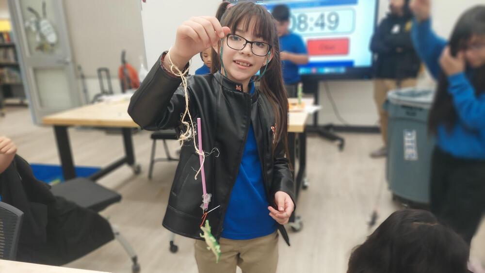 A student wearing glasses, a black jacket, and a blue shirt holds up a handmade classroom project consisting of a pink straw, string, and a small green figure. Other students, desks, and a digital display are visible in the background of the classroom.