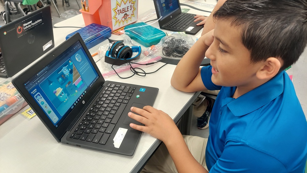 A student in a blue polo shirt sits at a classroom table using an HP Chromebook. The Chromebook displays an educational coding or computer-based learning activity. Several other Chromebooks, headphones, pencil cases, and classroom supplies are visible on the tables around the student.