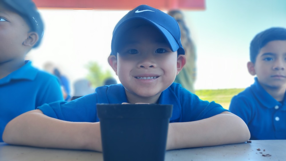Photo of a smiling young child seated at a table outdoors or under a covered area during a school activity. The child is wearing a blue collared shirt and a dark baseball cap and is leaning forward with arms resting on the table. A small black planting pot sits in the foreground. Other children in blue shirts are seated nearby, with the background softly blurred.