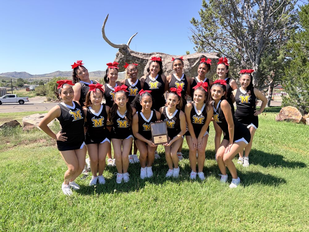 A cheerleading team of 14 members wearing matching black sleeveless uniforms with gold "M" logos and red hair bows poses outdoors on a grassy lawn. Two cheerleaders in the center hold up a wooden award plaque in front of a large longhorn sculpture.