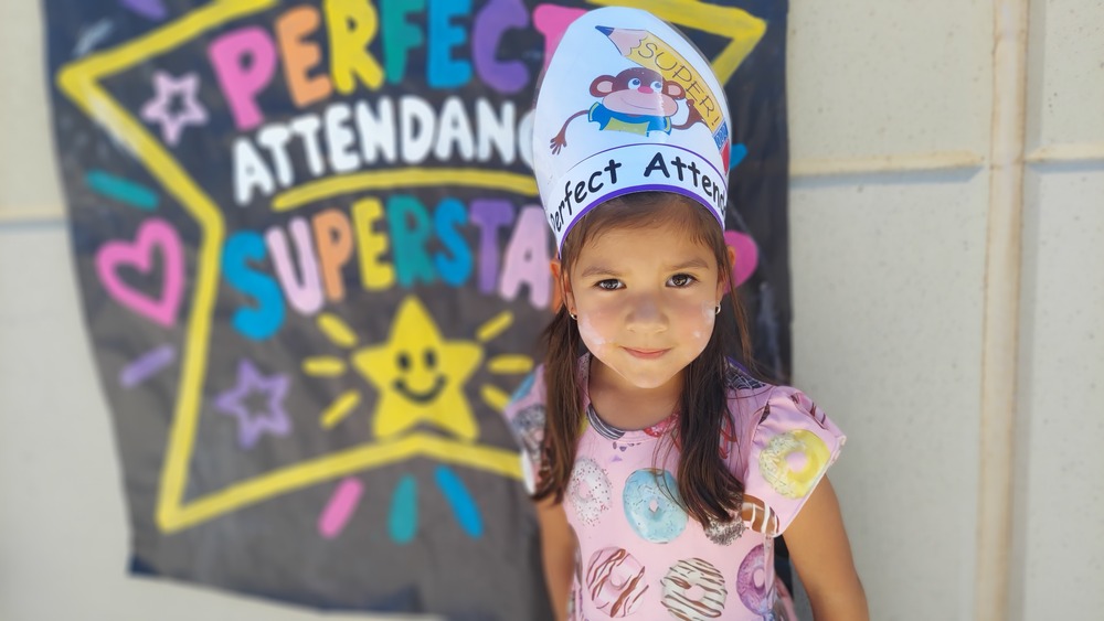 A young girl stands outdoors in front of a colorful chalkboard-style display celebrating “Perfect Attendance Superstars.” She is smiling at the camera and wearing a paper crown decorated with a cartoon character and the words “Perfect Attendance.” The girl has long brown hair and is wearing a pink shirt decorated with colorful donut designs. The background features a large yellow star outline, colorful lettering, stars, hearts, and a smiling star illustration.