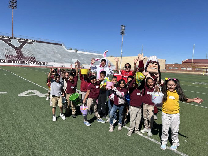 A group of excited elementary school children and adults posing together on the Ysleta High School football field on a sunny day. Many kids are holding colorful Easter buckets and waving at the camera. The group includes a person in a white bunny costume, another in a large Ysleta Indian mascot head, and chaperones. They are standing in front of the stadium bleachers under a clear blue sky.