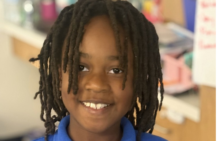 Portrait photo of a smiling young child indoors wearing a bright blue collared shirt. The child has shoulder-length dreadlocks and is looking toward the camera. The background appears to be a classroom or school setting with shelves and supplies softly blurred behind them.