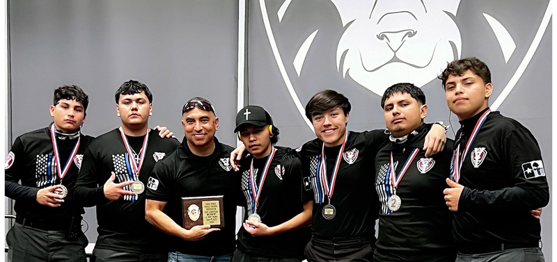 Group photo of the Ysleta High School Tribe Watch / Law Enforcement SWAT Team celebrating their 2nd place finish at the 2026 Texas Public Service Teachers Association (TXPTSA) State Competition in the Emergency Response Team (ERT / 6-Man Team) category. Seven team members, wearing black tactical uniforms with American flag and cross patches, medals around their necks, stand together smiling and with arms around each other. In the center, a coach or advisor in a black polo shirt holds a wooden plaque. Behind them is a large gray backdrop with a lion mascot logo. In the foreground, a polished wooden award plaque is displayed on the floor, engraved with: "TEXAS PUBLIC SERVICE TEACHERS ASSOCIATION STATE COMPETITION ERT (6 MAN TEAM) 2nd PLACE 2026," featuring the association's seal with American flag, badge, and medical symbols. The team poses proudly in an indoor venue with carpeted floor.
