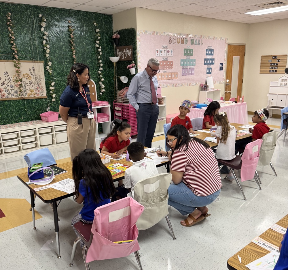 A group of young students work on assignments at a classroom table while two adults observe and one adult assists a student.