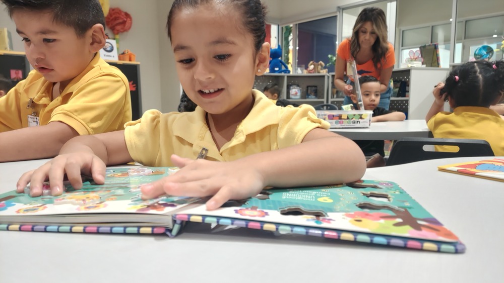 A Parkland Pre-K student sits at a table and explores a colorful activity book as part of a literacy center. The student uses both hands to interact with the pages and pictures. Other students are engaged in learning activities at tables in the background, with an adult assisting them. Books, classroom materials, and learning resources are visible throughout the classroom.