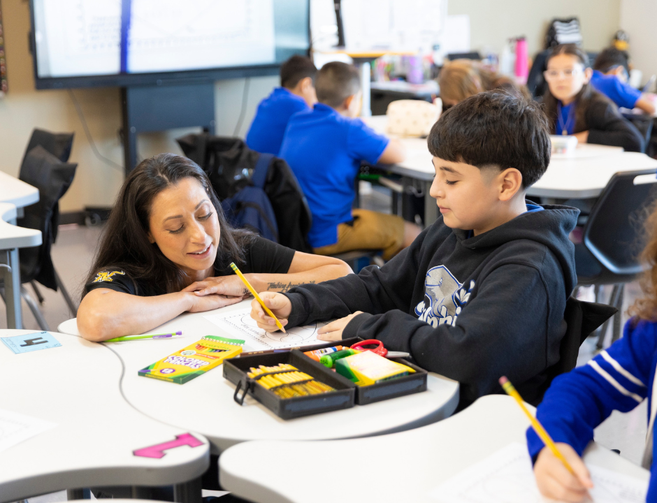 A teacher leans over to help a student who is drawing at his desk. Crayons, pencils, and papers are spread out. Other students work in the background.