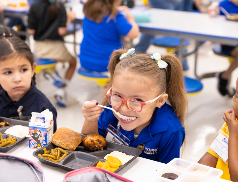 Three young girls sit at a cafeteria table eating lunch. One girl in red glasses smiles while taking a bite of food, and the others look at their trays with milk cartons, bread, vegetables, and dessert.