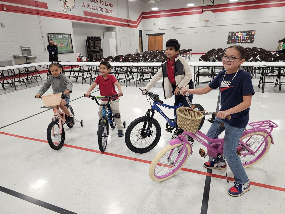 Moving from left to right. Four students, one Kinder girl, one 1st grade boy, a 5th grade boy and a 3rd grade girl. sitting proudly on the bikes they won for the perfect attendance