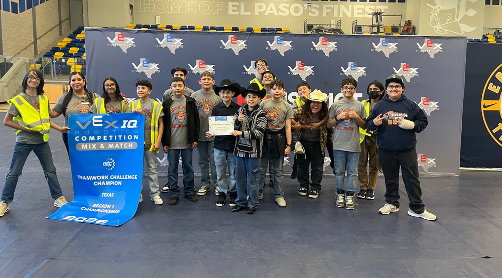 it shows students taking a picture with their robotics coach infront of the blue banner that confirms they won team work champion award while standing in front of the camera. A total of 17 individuals are in the picture including the coach