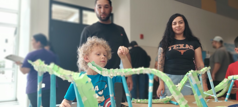 A young child with curly blond hair concentrates on placing a marble on  a handmade paper roller-coaster-style structure on a table during a community or classroom activity. Two adults stand behind the child watching supportively, while several other people are blurred in the background of the indoor space. The colorful structure is made from green-and-white paper strips and blue supports, extending across the foreground.