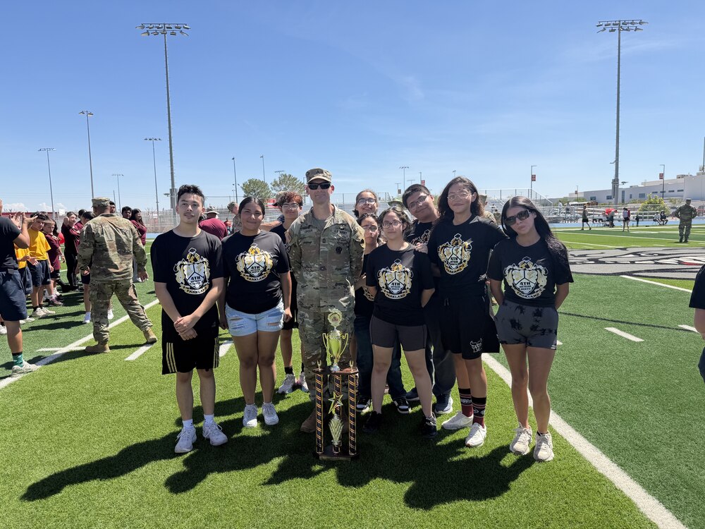A group of seven students wearing matching black Parkland 4th Battalion JROTC t-shirts pose on a green turf athletic field under a clear blue sky. A U.S. Army officer in camouflage stands in the center of the group, and a large multi-tiered gold trophy sits on the ground in front of them. In the background, other spectators and light poles surround the field.