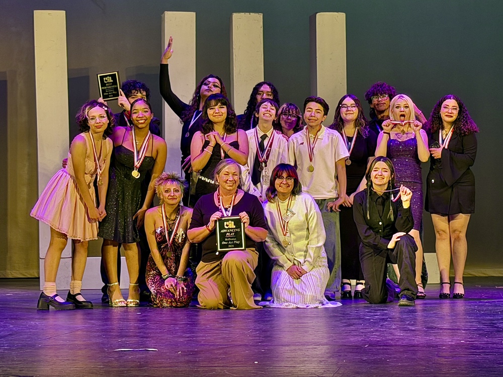 A group of smiling theater students and two adults pose on stage wearing medals. Two women in front kneel while holding a UIL "Advancing Play Bi-District One Act Play 2026" plaque. Another "All-Star Performers" plaque is visible in the back. Purple stage lighting.