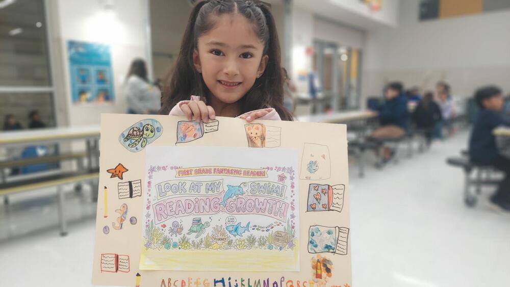 A smiling young girl stands in a school cafeteria, holding up a colorful, hand-decorated reading project. The poster features drawings of books, sea animals, and the alphabet, along with the phrases “First Grade Fantastic Reader!” and “Look at My Reading Growth!” Other students sit at tables in the softly blurred background.