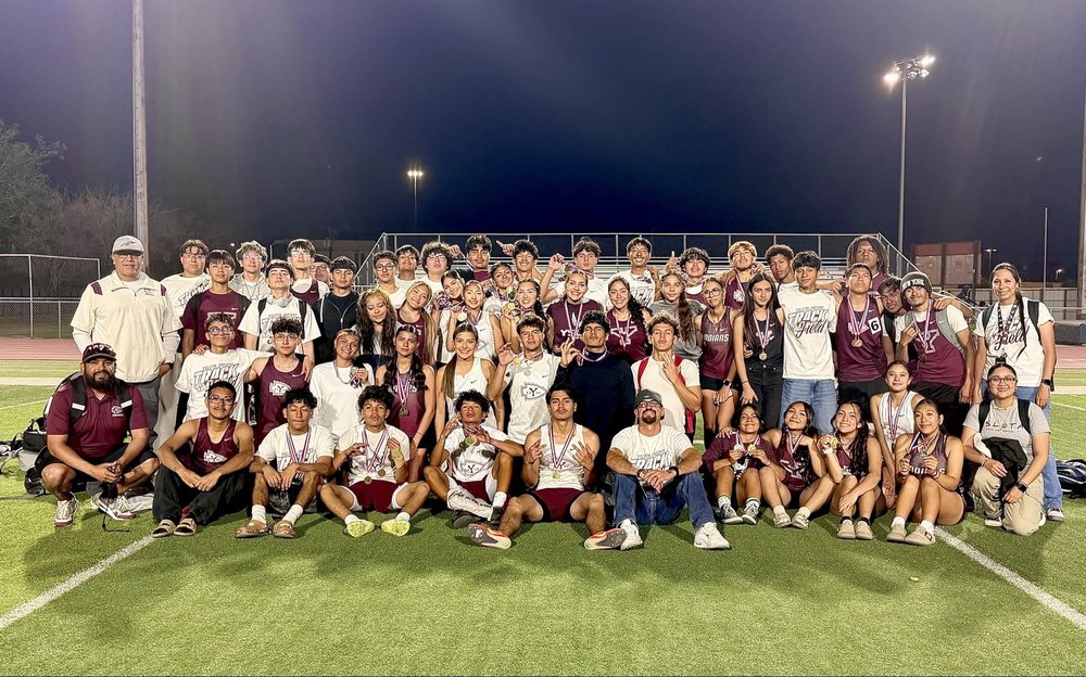A large group of Ysleta High School track and field athletes posing for a team photo on the football field at night. Many students are wearing medals around their necks, with some making peace signs or thumbs-up gestures. Coaches and staff are included in the group, and the background shows stadium lights and bleachers under a dark sky.