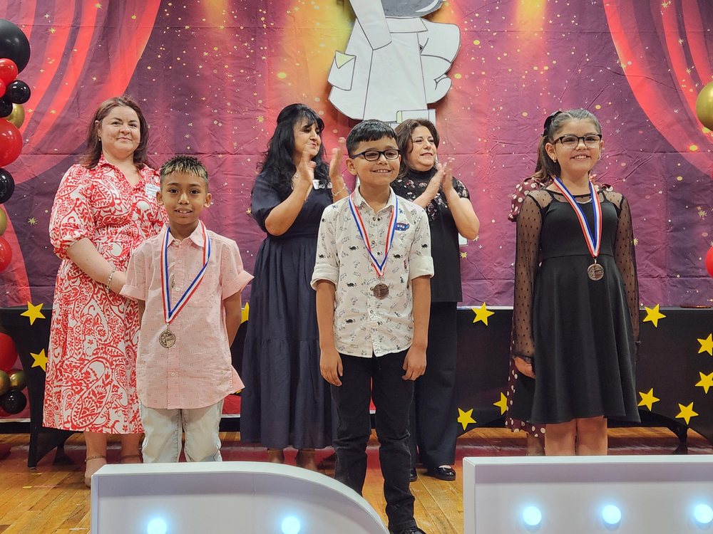 3 of our 2nd grade students standing on the stage after receiving medals with their teachers and principal standing in the back.