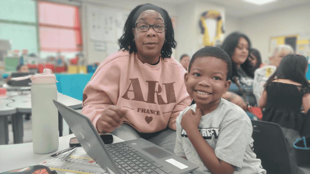 A classroom scene shows an adult seated beside a smiling elementary-aged student at a table. The student is seated in front of an open laptop and looks toward the camera. The adult wears glasses and a pink sweatshirt. Other students and adults are visible in the softly blurred background, along with classroom tables, supplies, and colorful decorations.