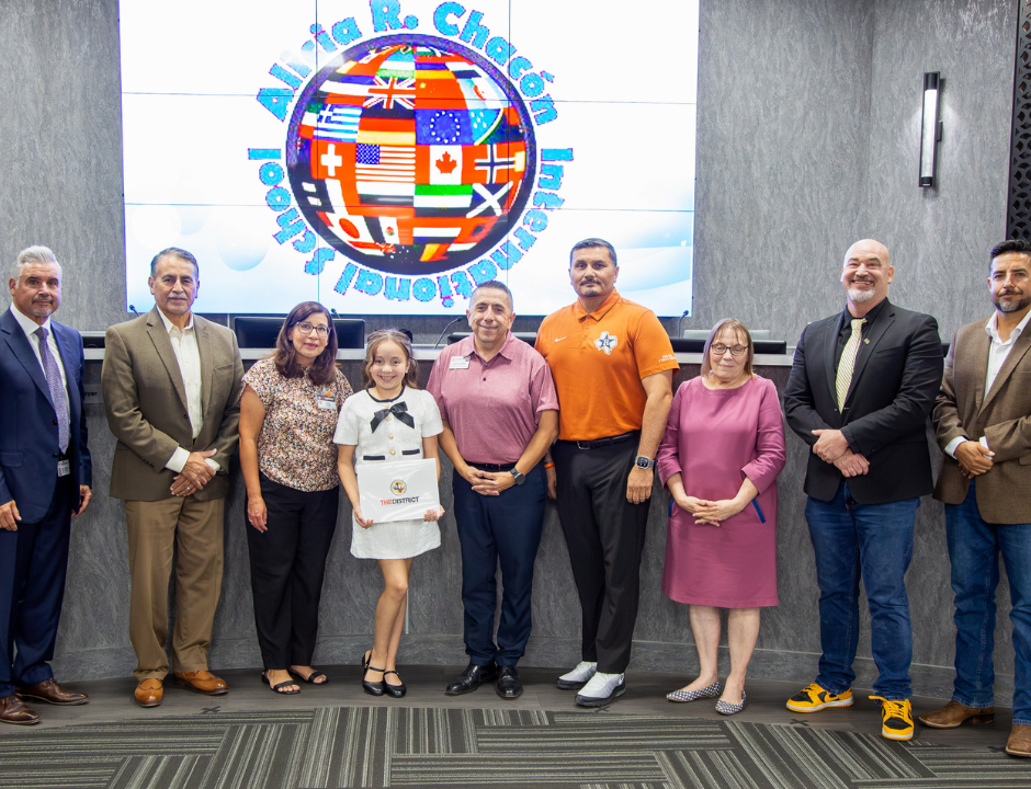 A group of adults and one child stand in a row, smiling at the camera. The child holds a certificate. Behind them is a screen displaying a globe with international flags and text: Alicia R. Chacón International School..