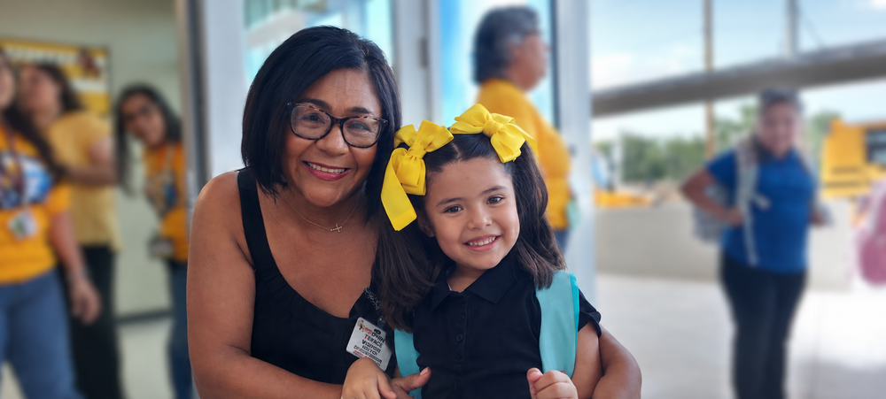 Mrs. Martinez is smiling as she welcomes a Pre-K student wearing a black uniform, turquoise backpack, and yellow hair bows inside the school.