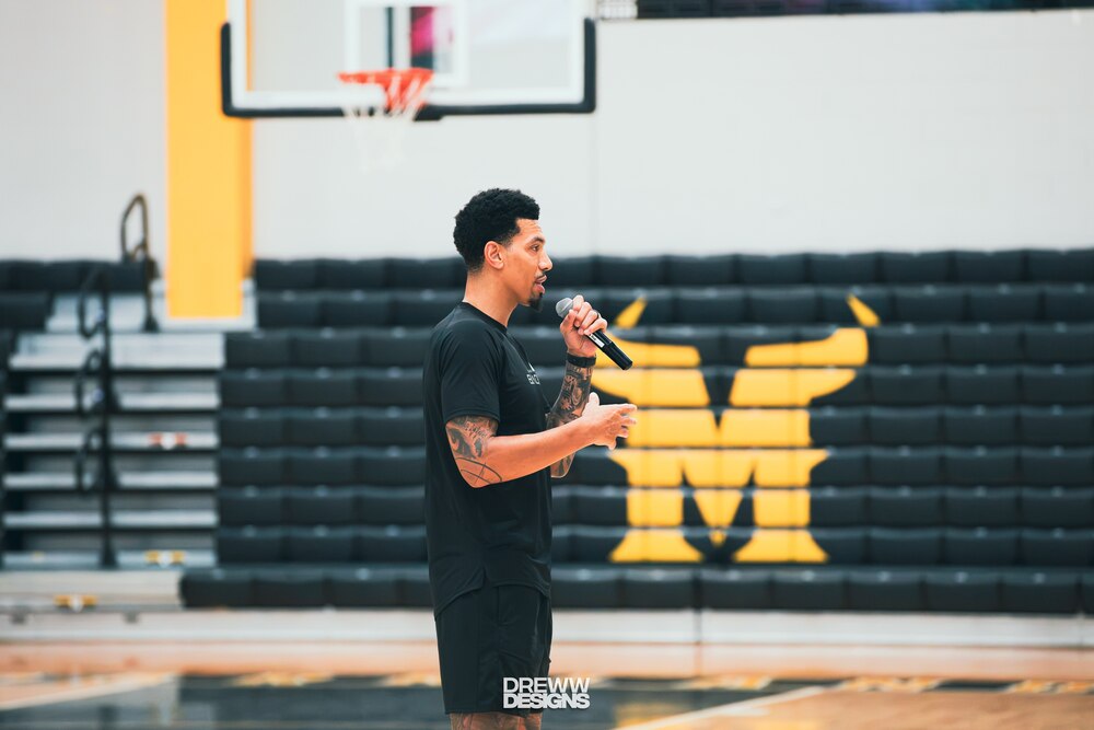 Danny Green speaking into a microphone on a basketball court inside a gymnasium with empty bleachers in the background.