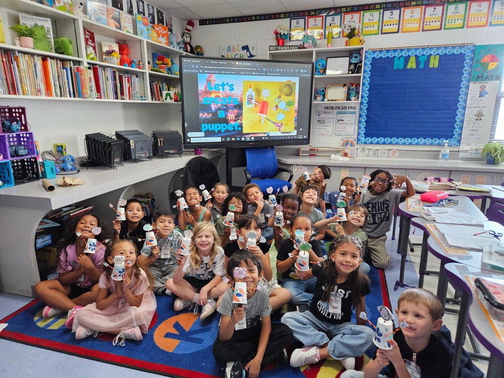 Students sitting on the carpet holding up puppets.