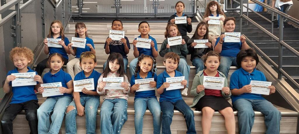A group of elementary students sit on indoor school stairs while holding “Dolphin of the Week” certificates. The students are arranged in several rows and smiling toward the camera. Most students wear blue school shirts with jeans or casual clothing. Metal railings and a modern school interior are visible in the background. The photo celebrates students being recognized for their positive achievements and hard work at school.