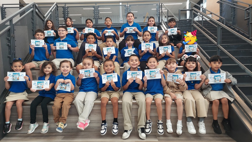 A group of elementary school students are seated together on indoor stairs for a group photo. The students are smiling and holding certificates recognizing them as “Dolphin of the Week.” Most students are wearing blue school shirts with khaki or neutral-colored bottoms. The photo is taken in a school stairway area with metal railings and a hallway visible in the background.