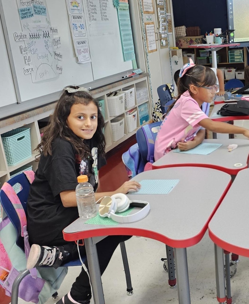 An elementary school girl sitting at her classroom desk smiles at the camera while another student works beside her.