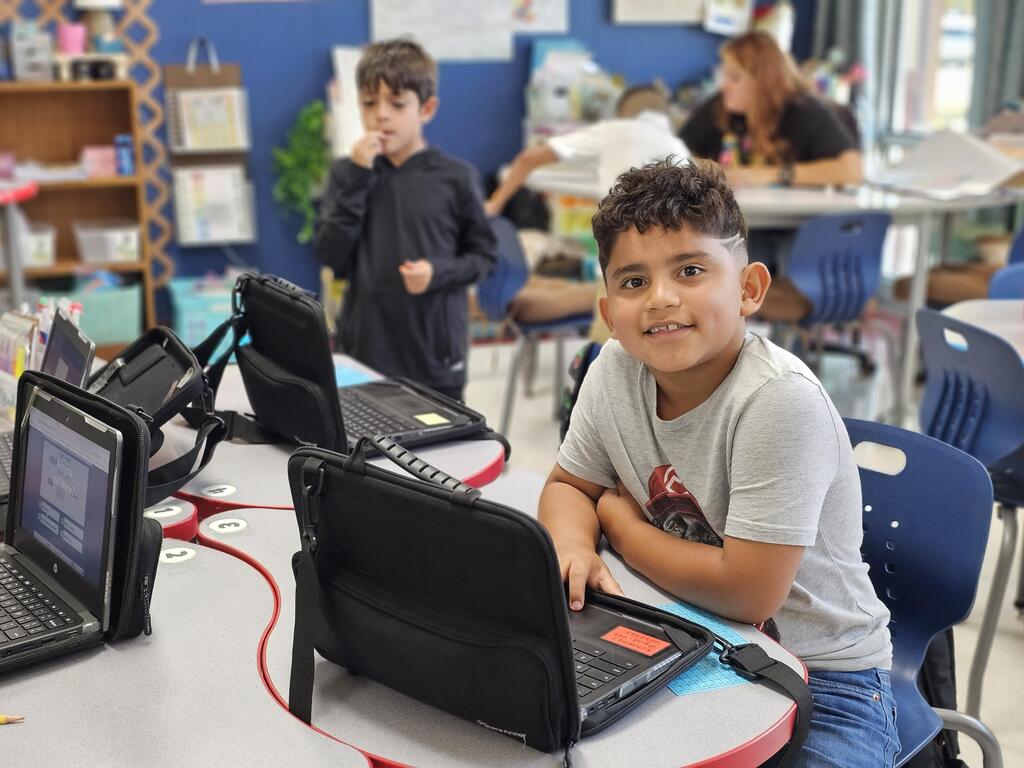 An elementary school boy smiles at the camera while using a laptop at his classroom desk.