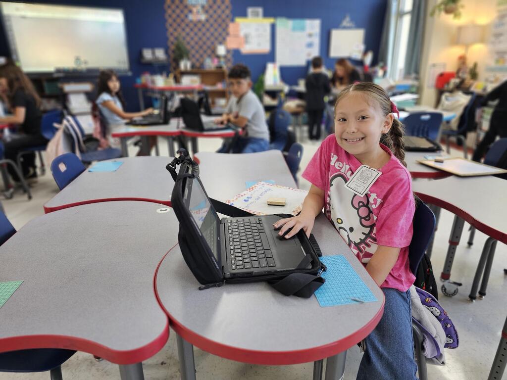 A young elementary school student wearing a pink shirt smiles at the camera while working on a laptop at her classroom desk.