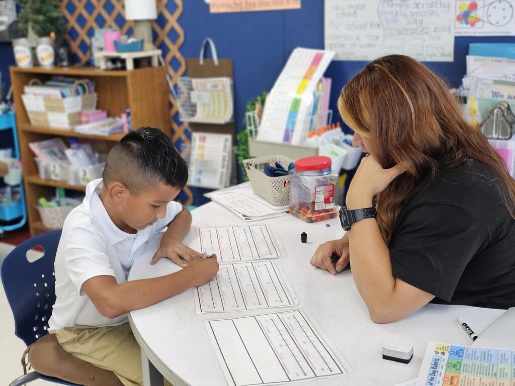 A teacher watches closely as a young male student works on a writing worksheet at a small table in a classroom.
