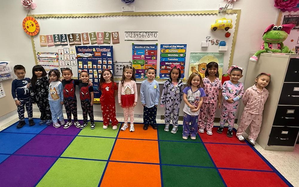 Thirteen young children lined up on a blue, green, orange, and red block-patterned rug, wearing diverse pajamas including superhero and dinosaur prints, standing in front of a decorated classroom whiteboard