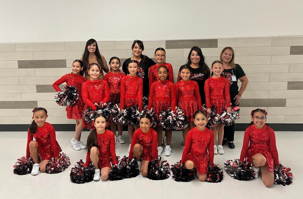 Dance team posing on a wooden stage. Young girls in red sequined dresses stand in front of teachers in black clothing. .