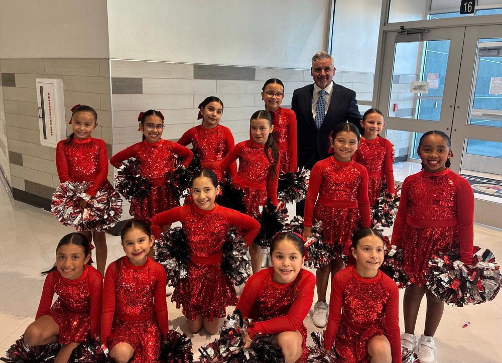 Young girls in red sequined dance outfits holding pom-poms pose with a man in a sui