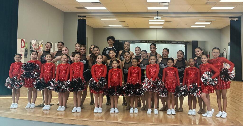 Dance team posing on a wooden stage. Young girls in red sequined dresses stand in front of older dancers in black.