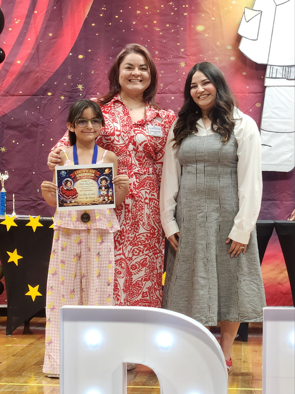 Third grade student holding her Explorador certificate and posing with her teacher and Ms. Ortiz on stage