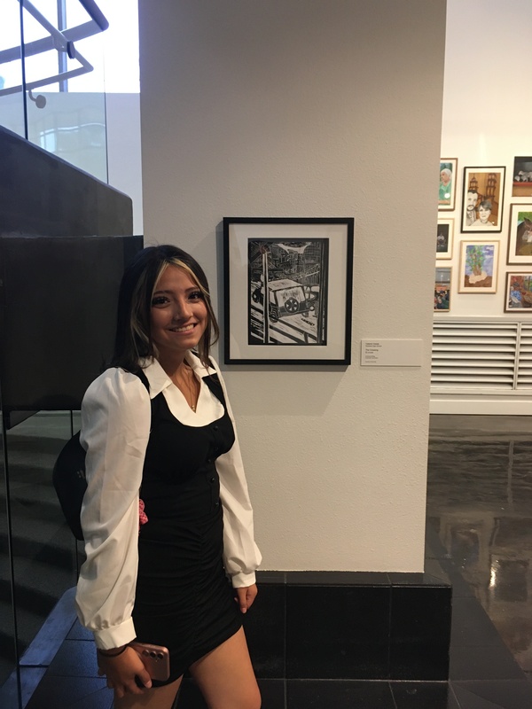Art student, Valerie Vela, stands near her displayed artwork high-contrast black-and-white print titled "El cruce / The Crossing."