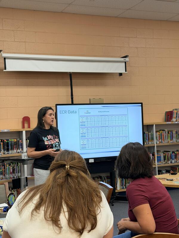 A female presenter stands in front of a digital screen displaying "ECR Data" charts, addressing two women seated in the foreground inside the Presa Elementary library.
