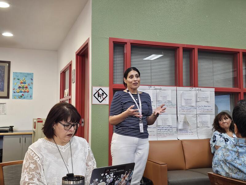 New principal Ms. Holguin stands and speaks with expressive hand gestures during a meeting in the Presa Elementary library, with whiteboards and colleagues seated nearby.