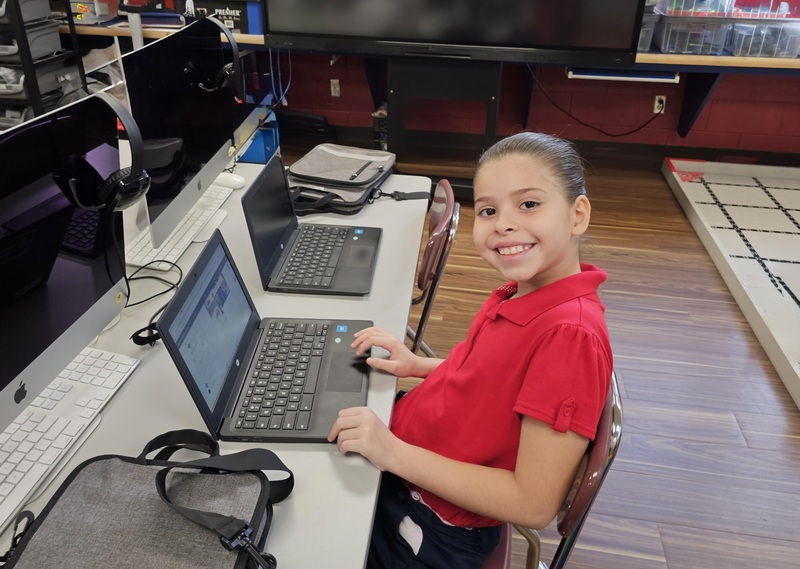 young female student in red shirt smiles at camera while sitting in front of her computer