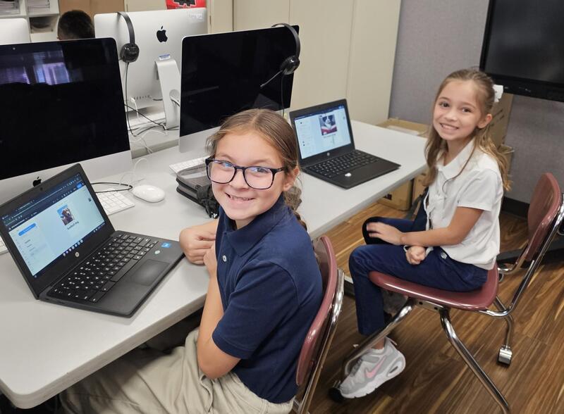 young female student in blue wearing glasses and another female student  with a white shirt and a white bow in her hair smile at camera while sitting in front of their computers