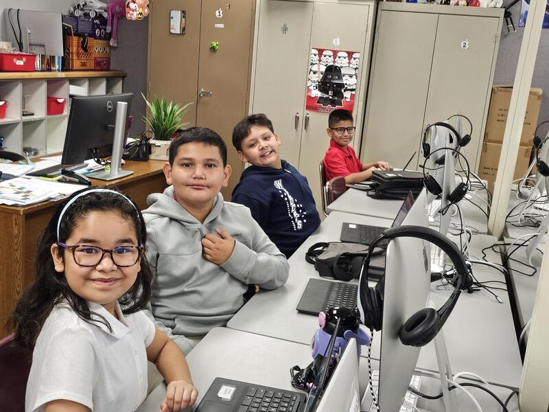 Four young elementary students smiling at the camera while seated in a row at computer lab workstations with laptops and desktop monitors.