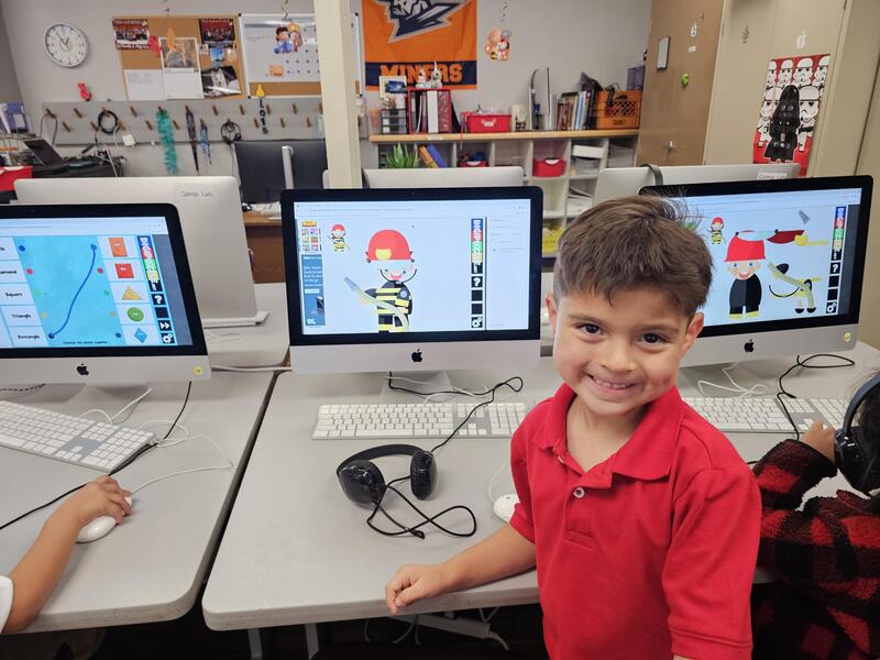Young male student stands in a red shirt front of large MAC desktop showing off his use of a mouse to move parts to recreate image of fireman