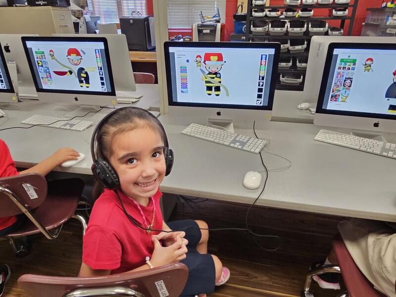 young female student in red shirt smiles at camera while sitting in front of the desktop computer with a fireman image on the screen 