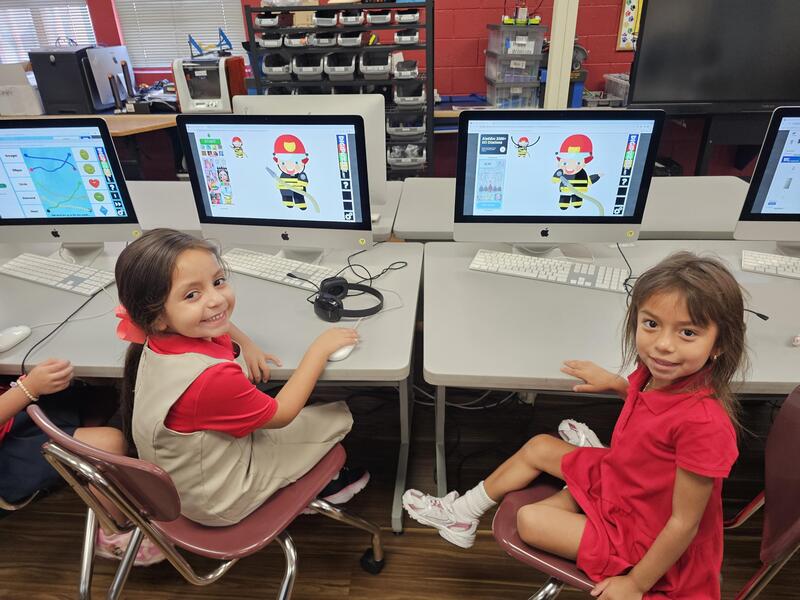 two young girls proudly smile at the camera while turned slightly in their chairs. Image of a fireman is on each screen
