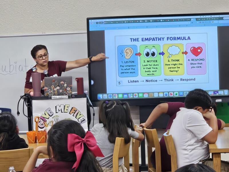 Mrs. Jordan teaches in front of the students in the library about Empathy. Students look on at the tables