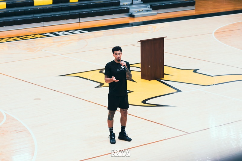 Danny Green holding microphone standing in middle of basketball court. Podium and the high school M logo are visible in the background.